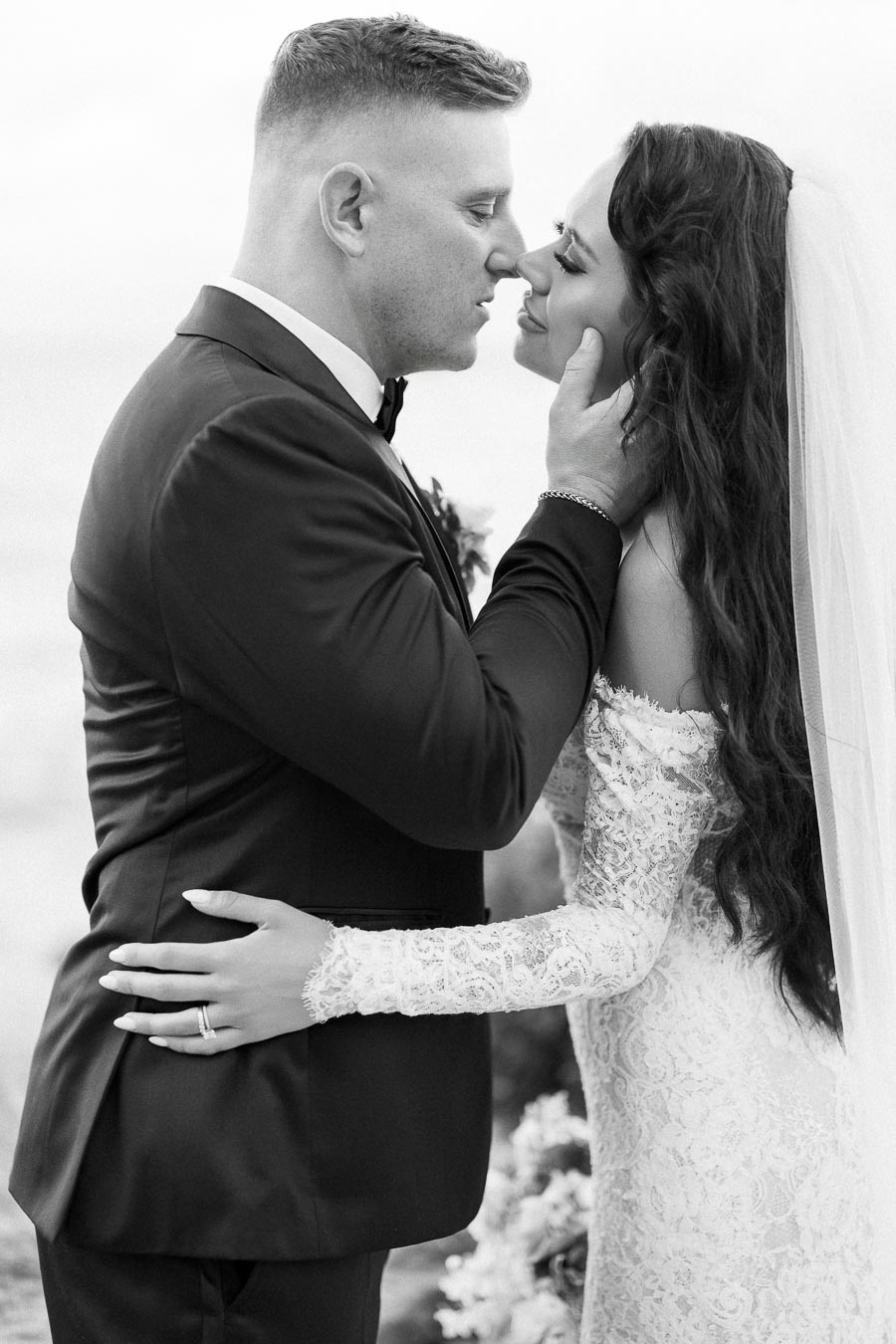 Black and white photo of a bride and groom sharing an intimate moment on their wedding day, the groom in a formal suit lovingly holds the bride's face, who wears a lace wedding dress and veil.