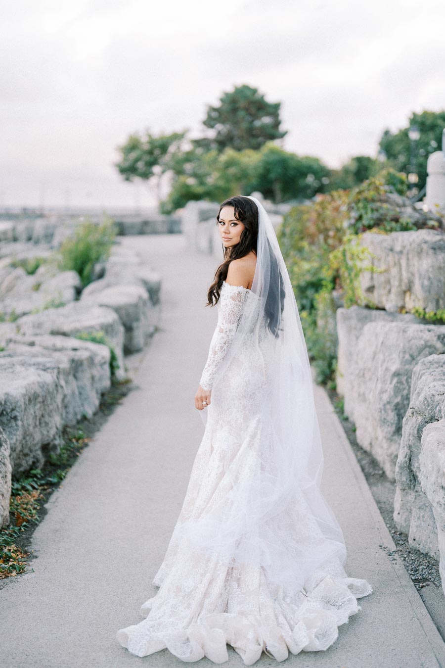 Bride in a lace wedding dress with long veil walks along a stone pathway, surrounded by greenery and rock formations.