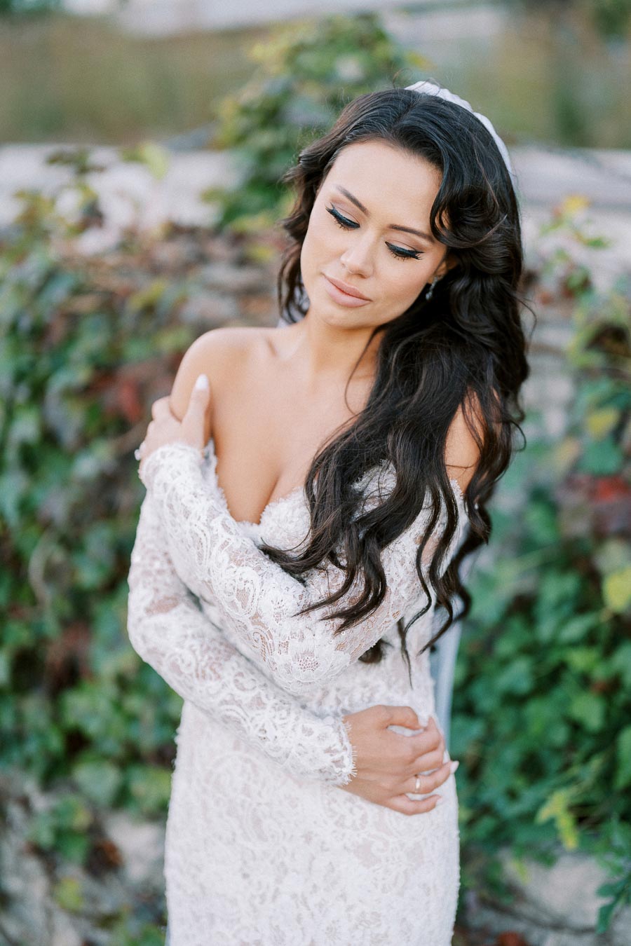 Female bride in elegant white lace wedding dress posing outdoors with floral background.