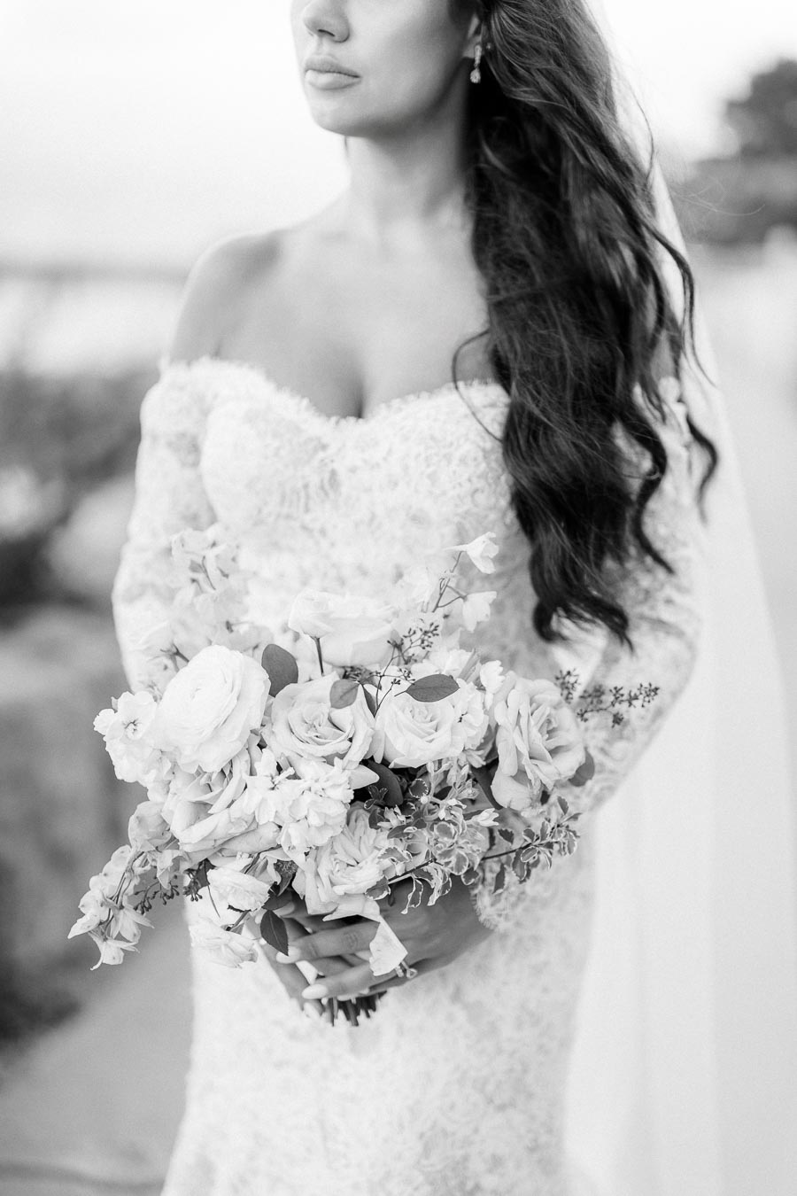 Black and white image of a bride holding a bouquet of white roses and flowers, wearing a lace off-shoulder wedding dress, showcasing elegant bridal fashion and romantic wedding themes.