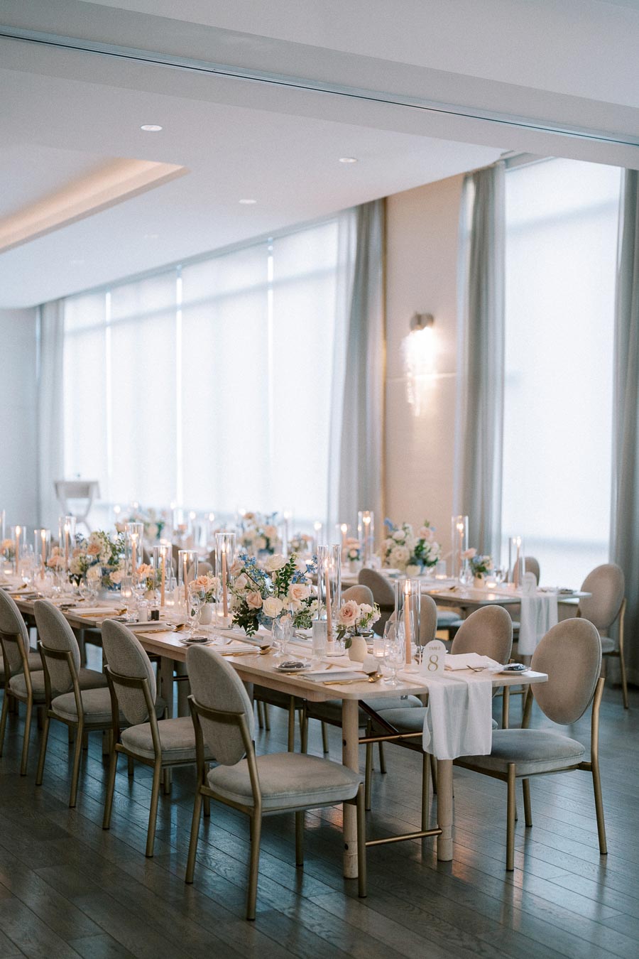 Elegant wedding reception setup featuring long tables adorned with floral centerpieces, candles, and fine tableware in a modern, sunlit room.
