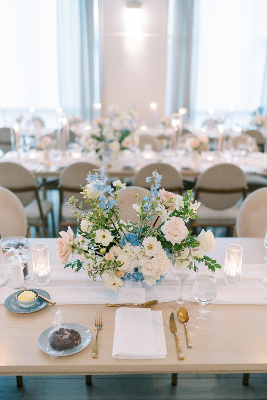 Elegant wedding table setting with a centerpiece of pastel flowers, including blue and white blooms, surrounded by fine dining cutlery, glassware, and candle holders.