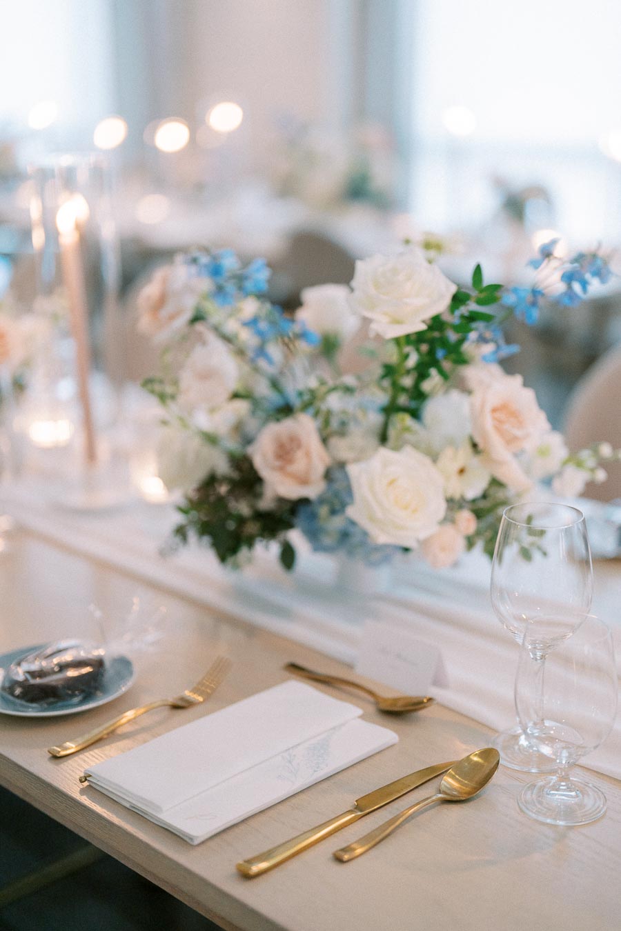 Elegant wedding table setting with gold cutlery, white napkins, wine glasses, and a centerpiece of white and pale pink roses with blue accents. Candles create a soft, romantic ambiance.