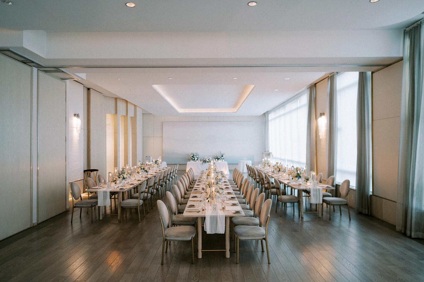 Elegant wedding reception venue with long tables set for dining, featuring soft lighting and floral centerpieces in a bright, modern banquet hall.