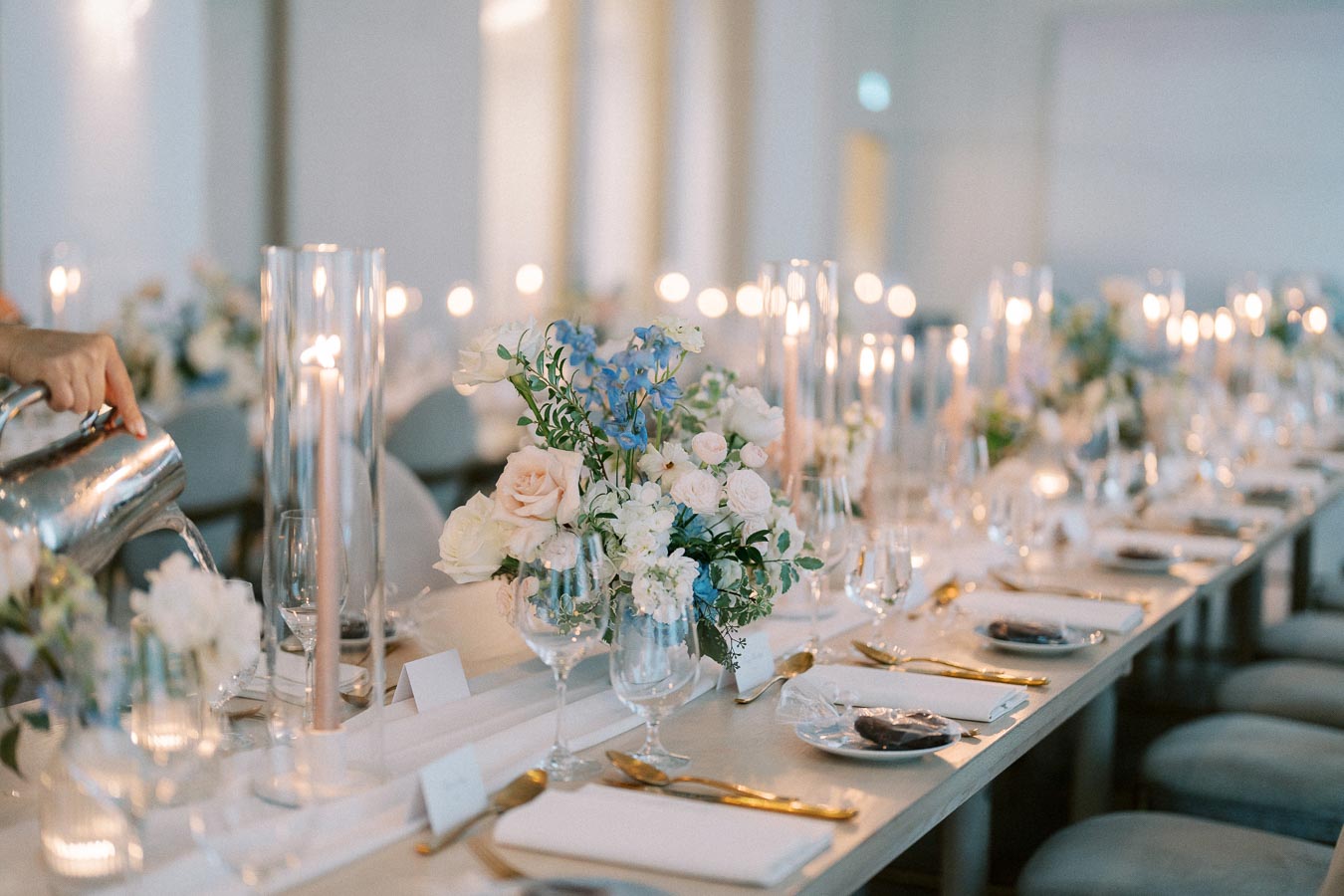 A beautifully set wedding reception table adorned with elegant flower arrangements of pastel roses and delphiniums, surrounded by flickering candlelight. A hand is pouring water from a jug into a glass, enhancing the serene and romantic atmosphere. Elegant tableware and gold flatware are neatly arranged, ready for guests.