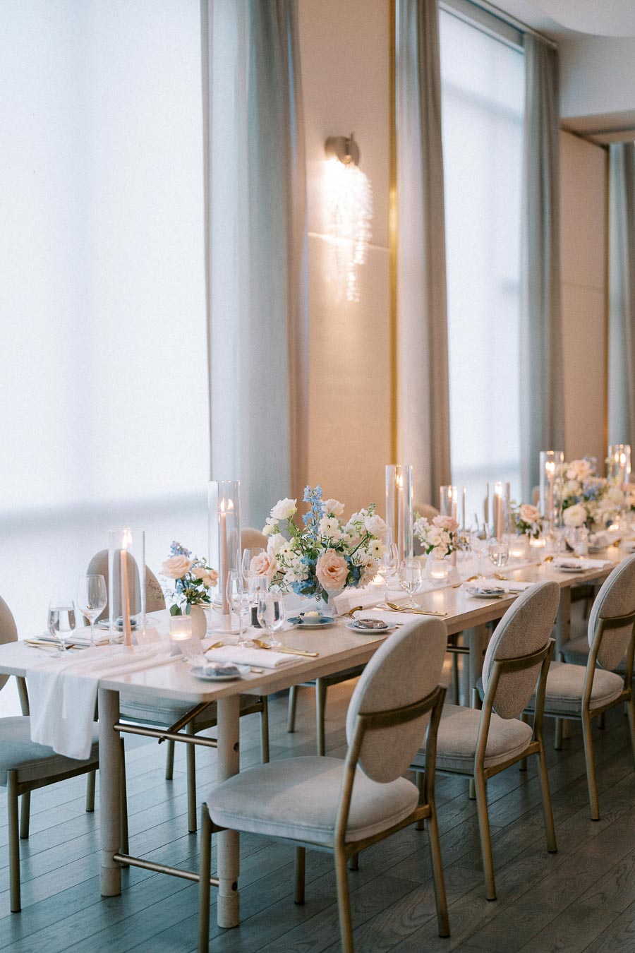 Elegant wedding reception table setting with soft blue and white floral arrangements, tall candles, and white chairs in a bright, sophisticated venue.
