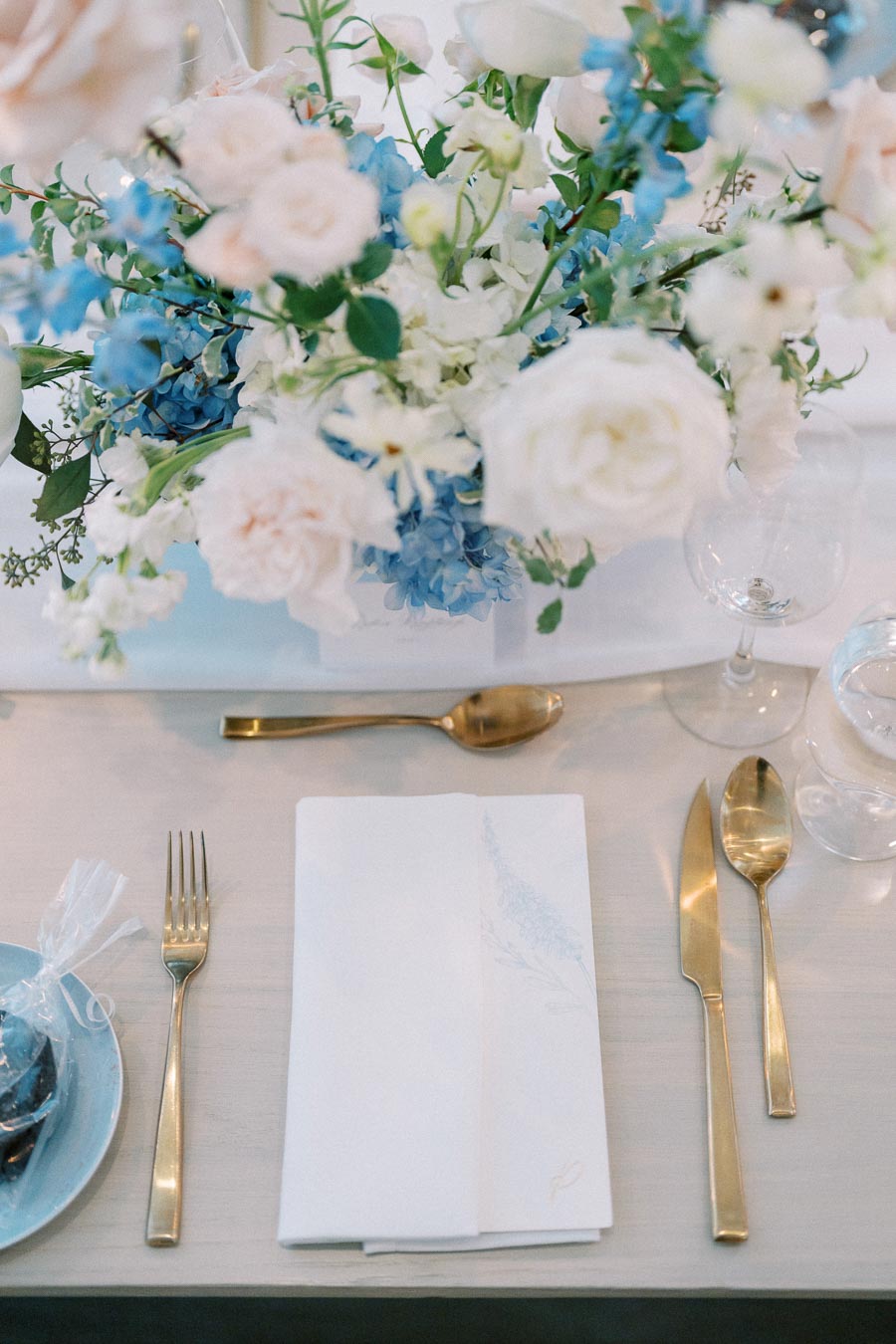 Elegant table setting featuring gold cutlery, a white and blue floral centerpiece with roses and hydrangeas, and a crisp white napkin for a sophisticated dining experience.