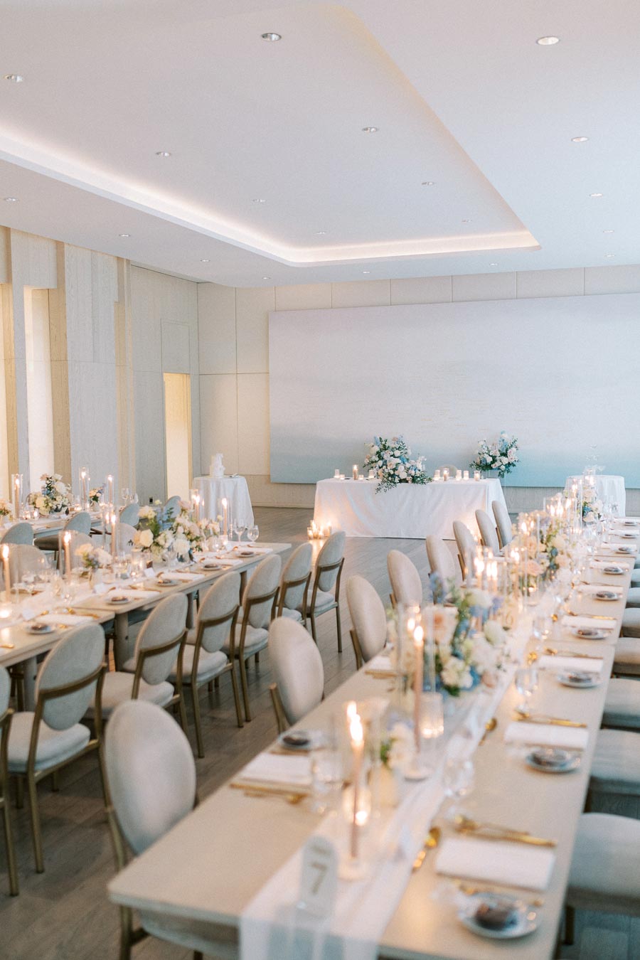 Elegant wedding reception setup in a modern banquet hall with long tables adorned with white floral centerpieces and candles.