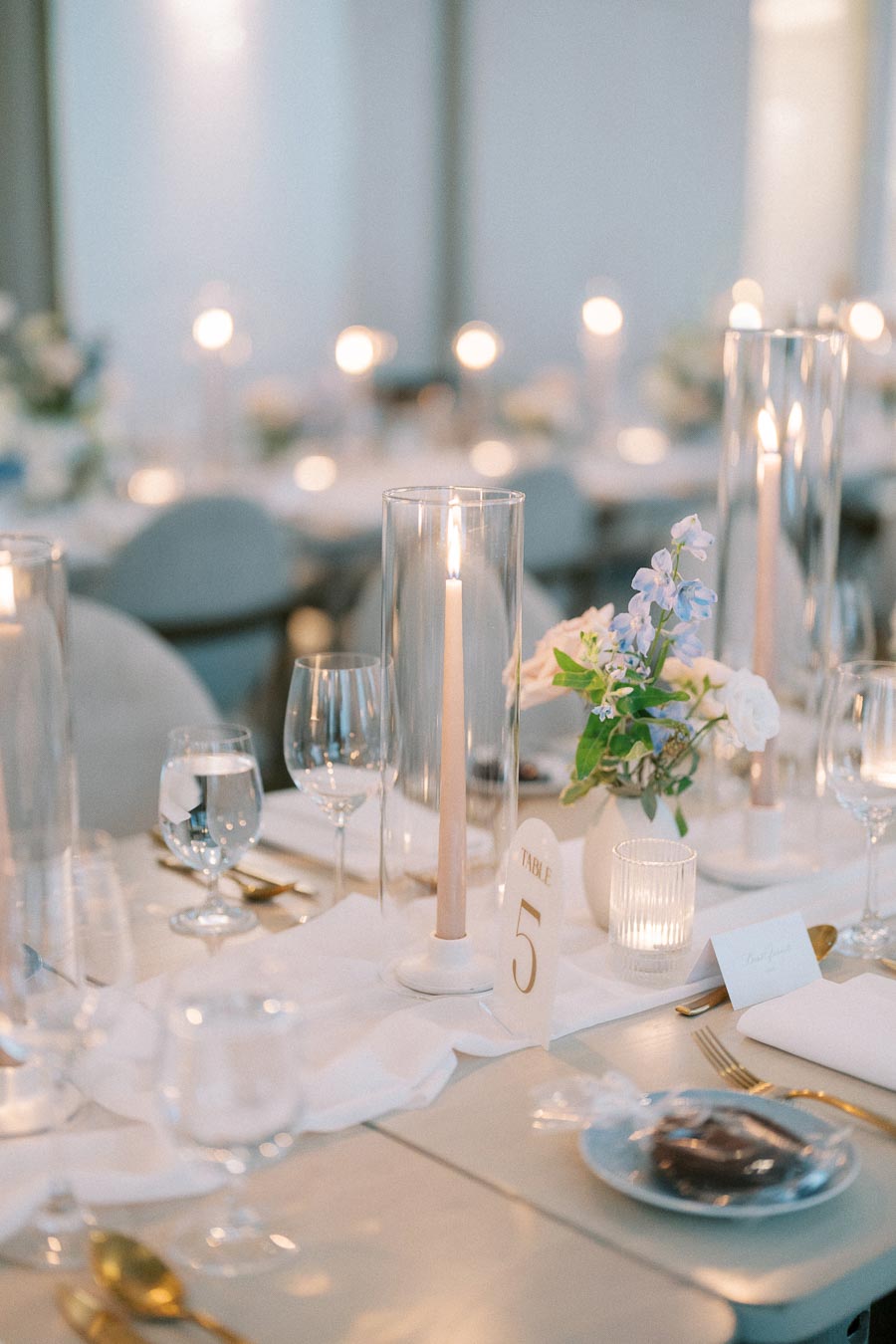 Elegant table setting with tall candles, glassware, and floral centerpiece at a wedding reception, featuring table number five in a softly lit banquet hall.