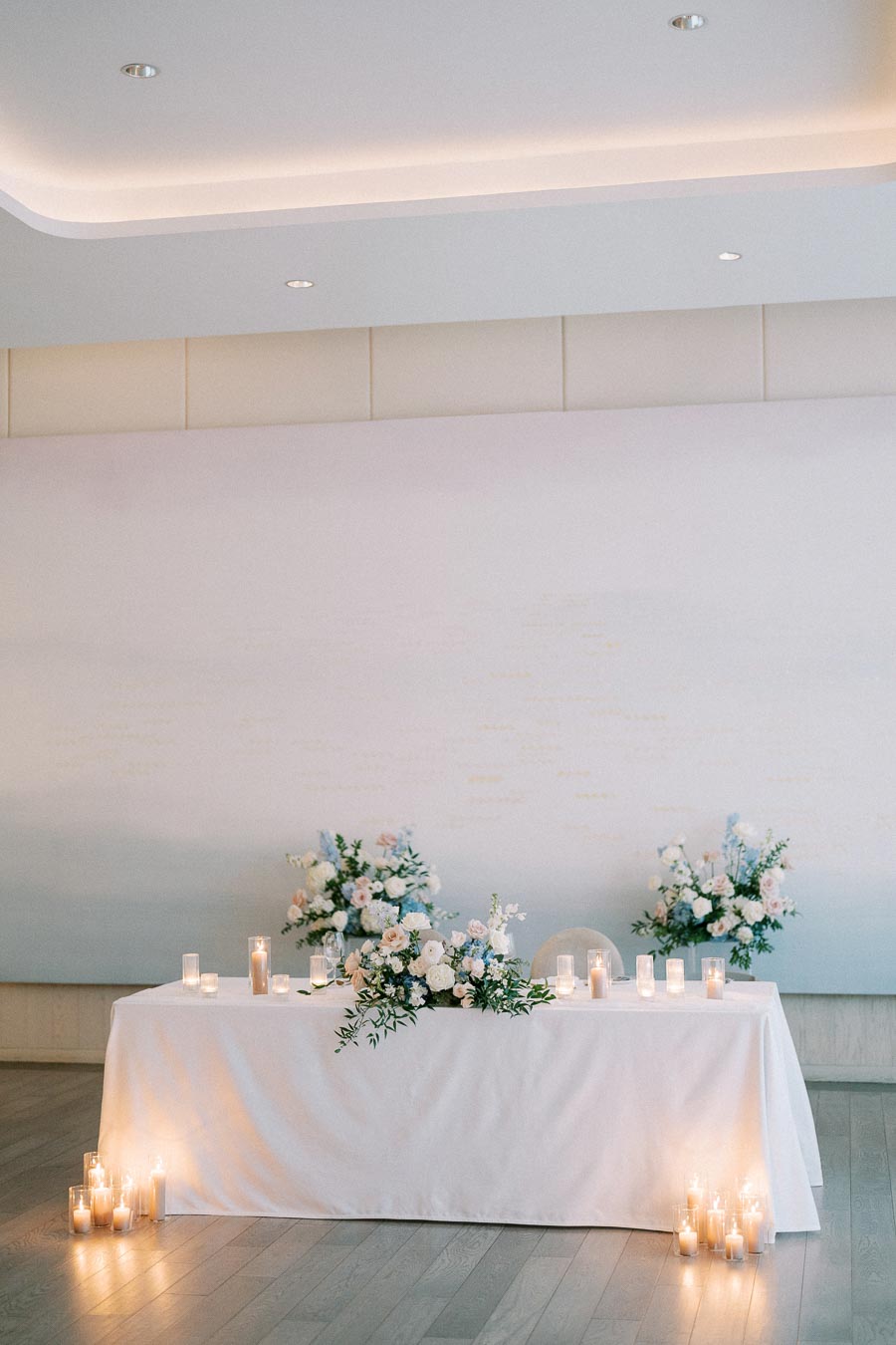 Elegant wedding reception table decorated with white and pastel floral arrangements, surrounded by glowing candles, set against a minimalist backdrop.