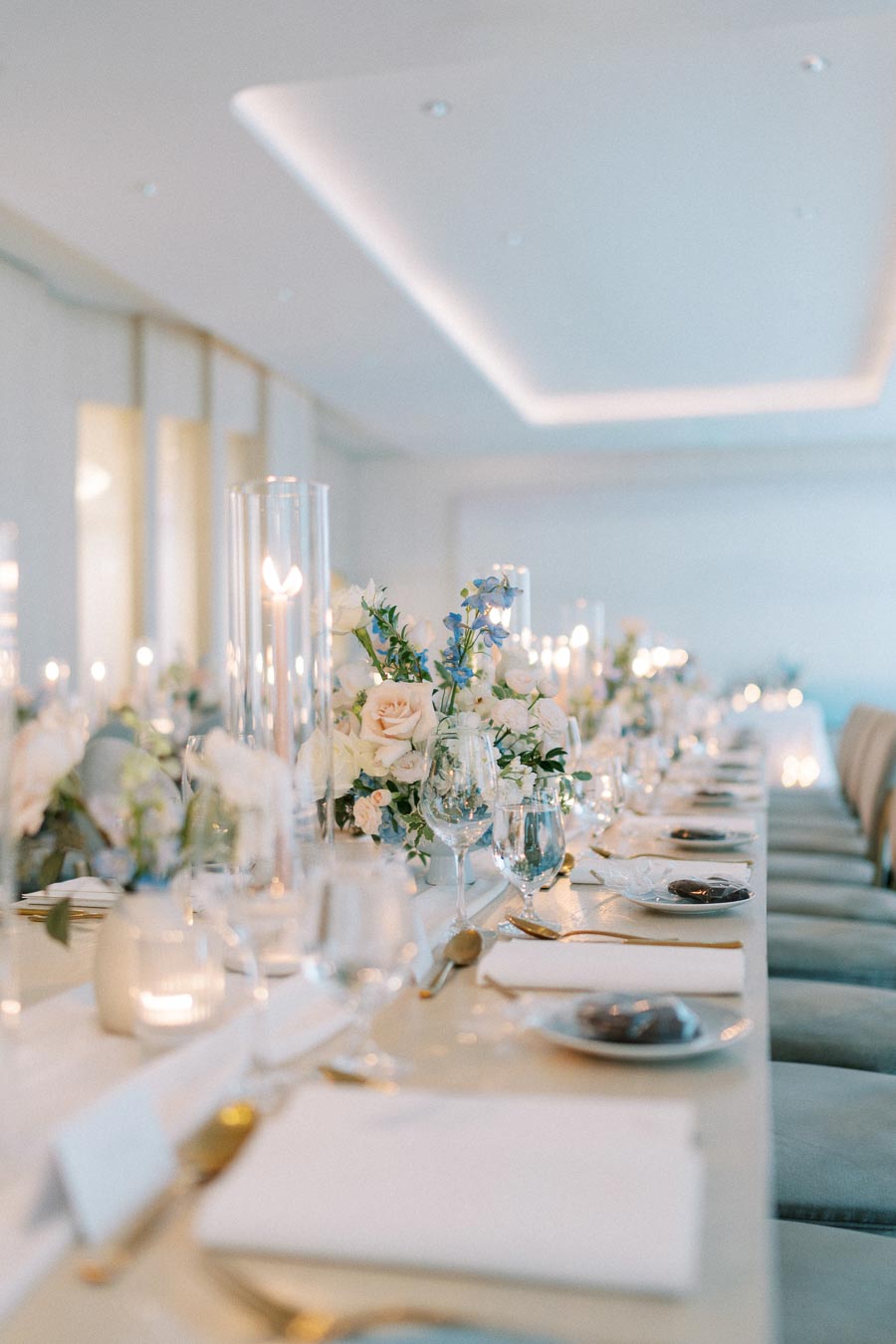 Elegant wedding reception table setting with white and pastel floral centerpieces, glassware, and candles in a bright, modern room.