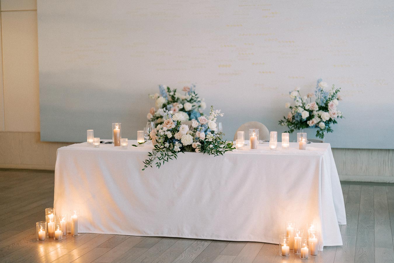 Elegant wedding table setting with white tablecloth, adorned with soft pastel floral arrangements and surrounded by glowing candles in glass holders, creating a romantic ambiance.
