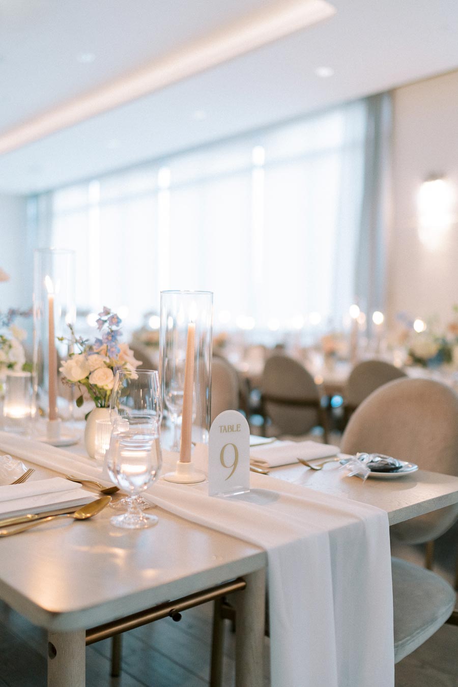 Elegant wedding reception table setting with white linen, floral centerpiece, number 9 table card, and candles in glass holders.