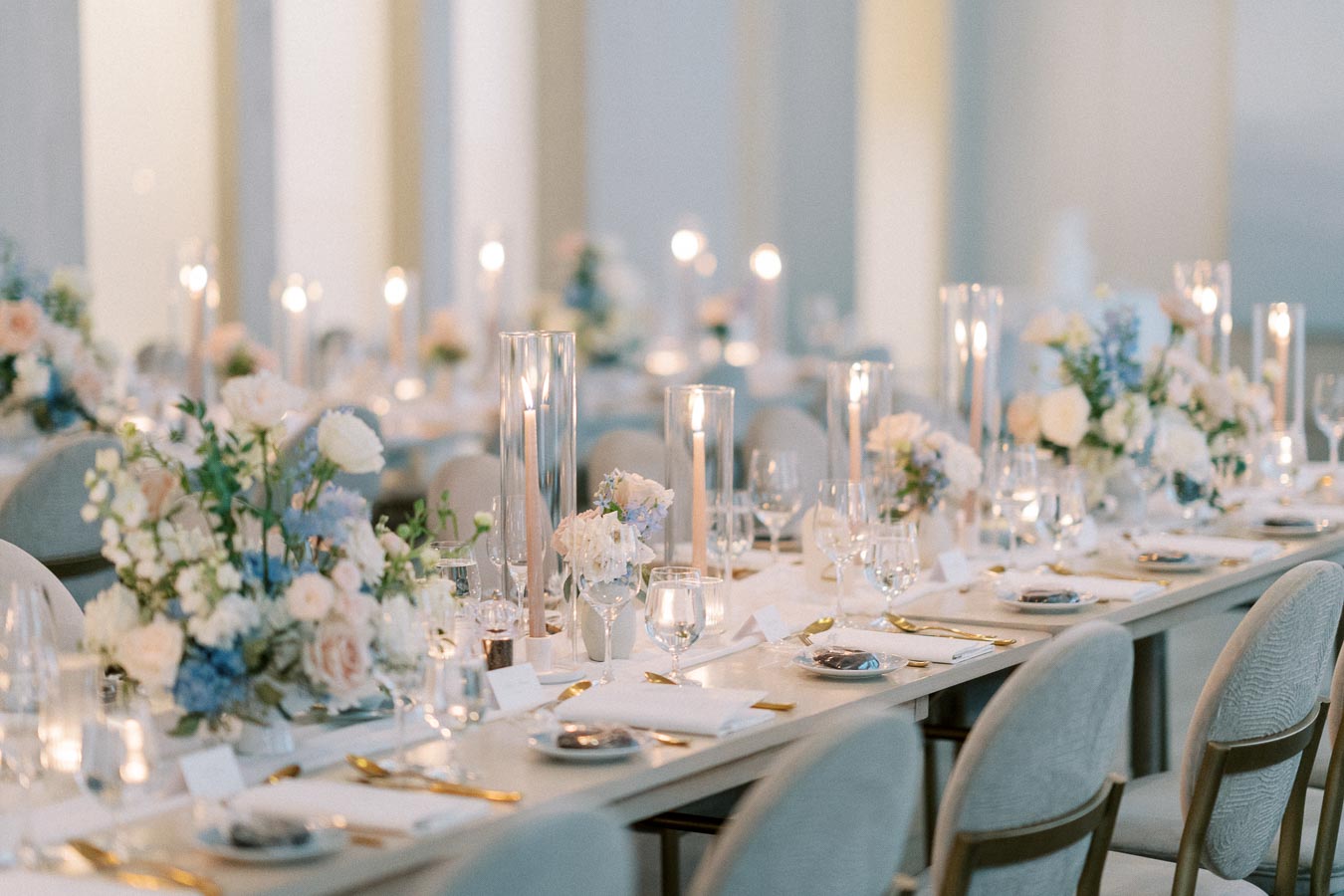 Elegant wedding reception table setup with candles, floral arrangements, and neatly arranged tableware in a softly lit venue.