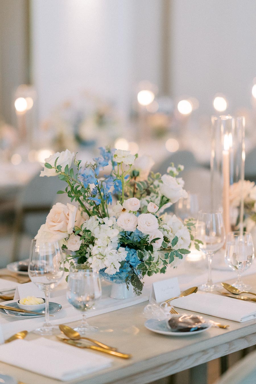 Elegant wedding table setting with a centerpiece of lush pastel roses, blue hydrangeas, and white lisianthus, surrounded by candlelit ambience and fine dining utensils.