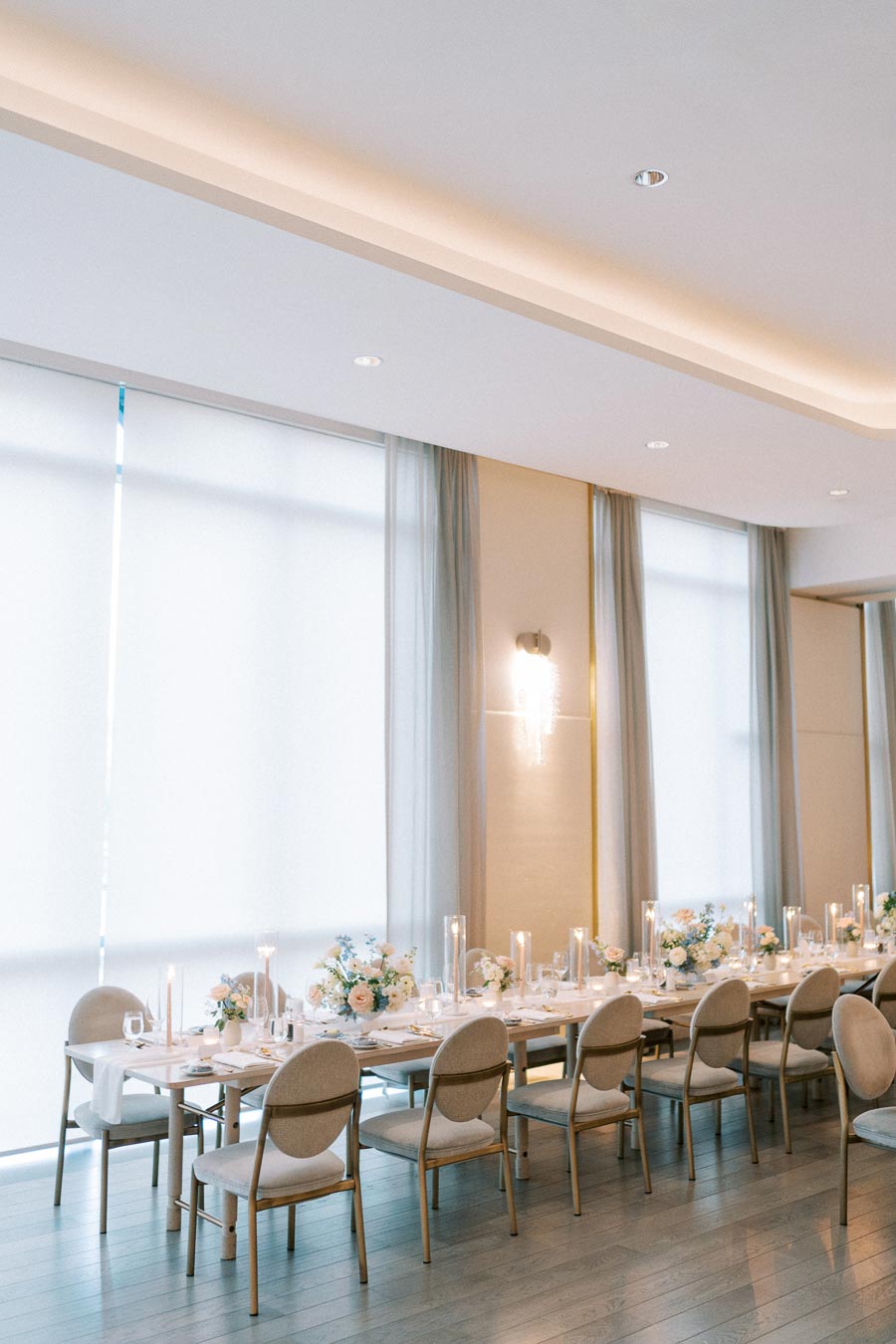 Elegant wedding reception setup with a long table adorned with floral centerpieces and candles, surrounded by beige chairs in a softly lit venue with tall windows.