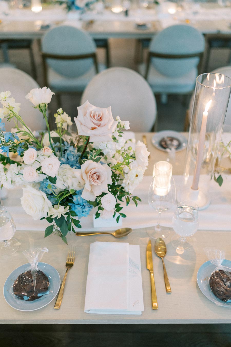 Elegant wedding table setting with a floral centerpiece featuring pastel roses and blue hydrangeas, surrounded by gold cutlery, glassware, and ambient candlelight.