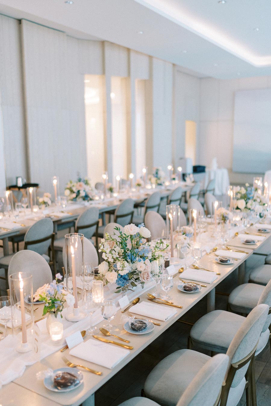 Elegant wedding reception table setting with white and blue floral arrangements, gold cutlery, and soft candlelight in a modern banquet hall.