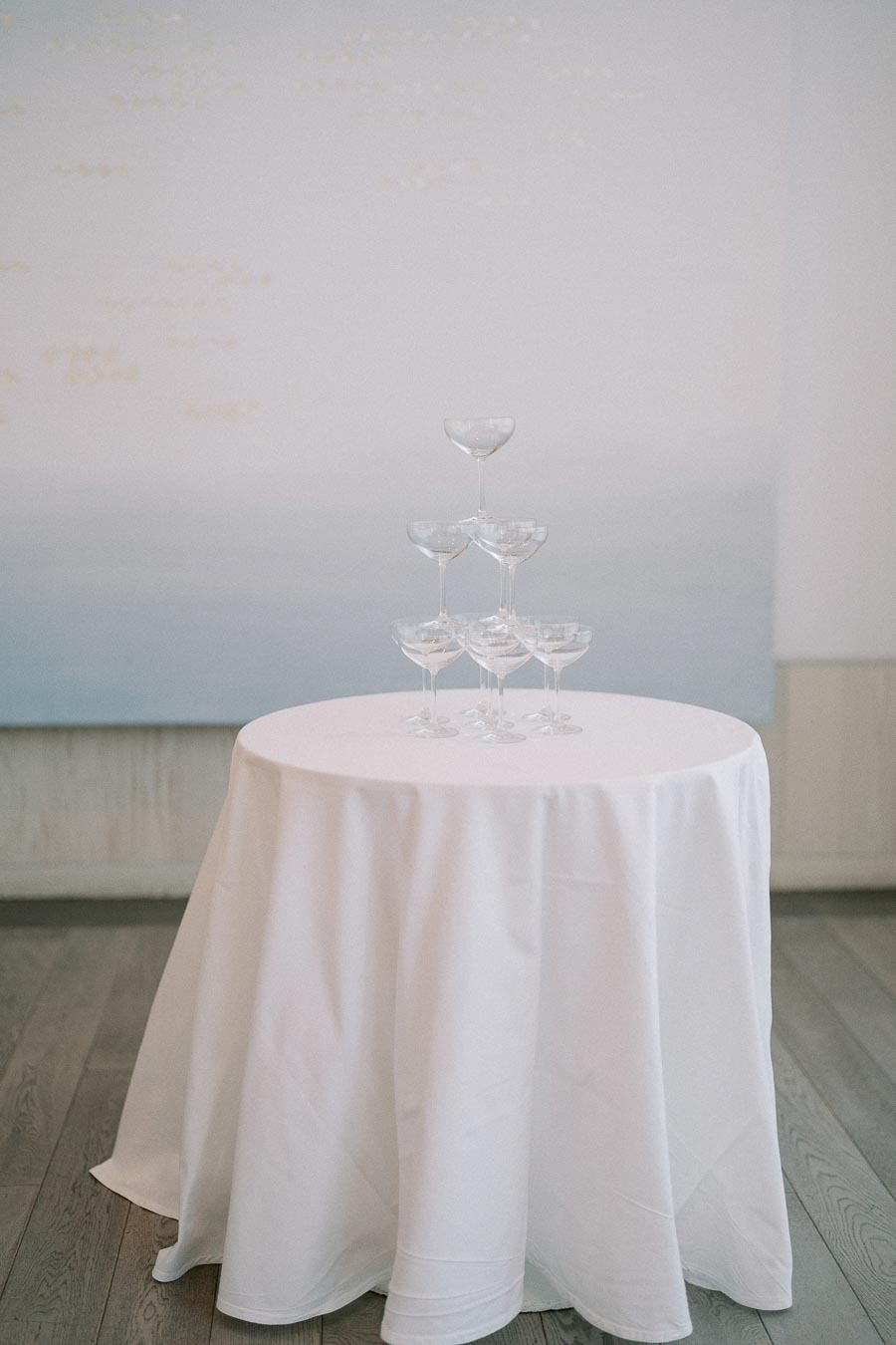 A pyramid of empty champagne glasses on a round table with a white tablecloth, set against a soft gradient background, perfect for elegant wedding receptions or sophisticated event decor.
