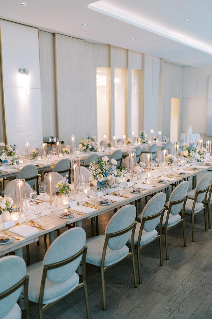 Elegant wedding reception setup with long dining tables, adorned with lush floral centerpieces, tall candles, and neatly arranged tableware in a modern venue with soft lighting.
