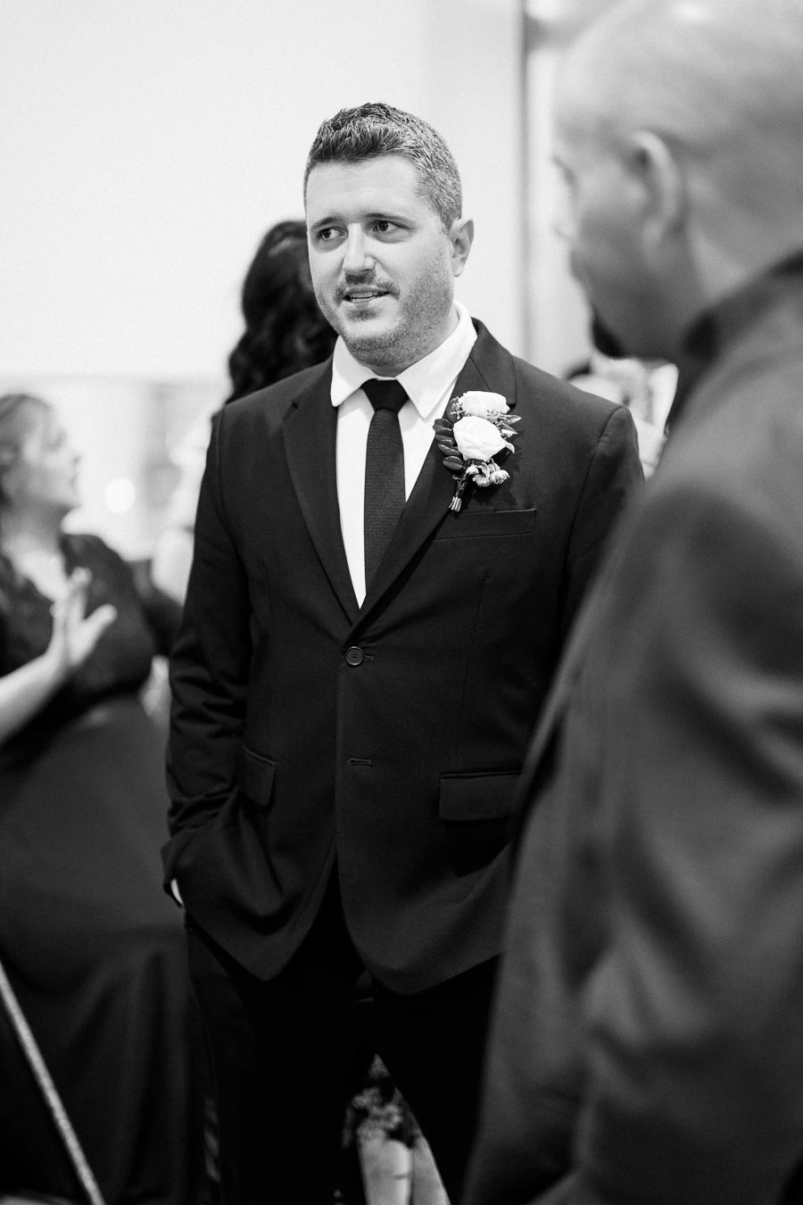 A black and white photo of a man in a suit with a boutonniere, standing with hands in pockets, engaged in conversation at a formal event.