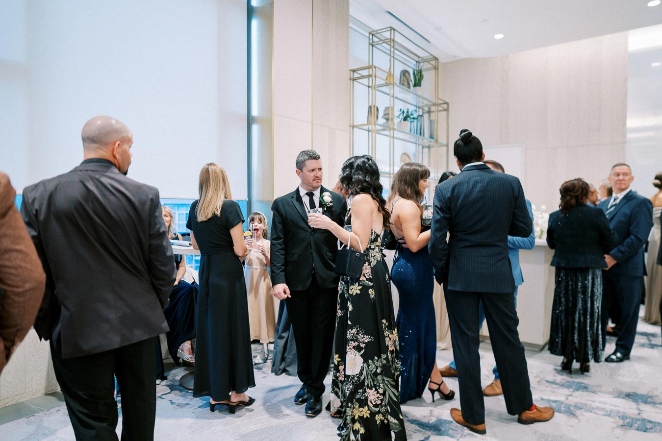 A group of elegantly dressed people mingling and socializing at a formal indoor event, with men in suits and women in gowns, set in a modern venue with decorative shelves and soft lighting.