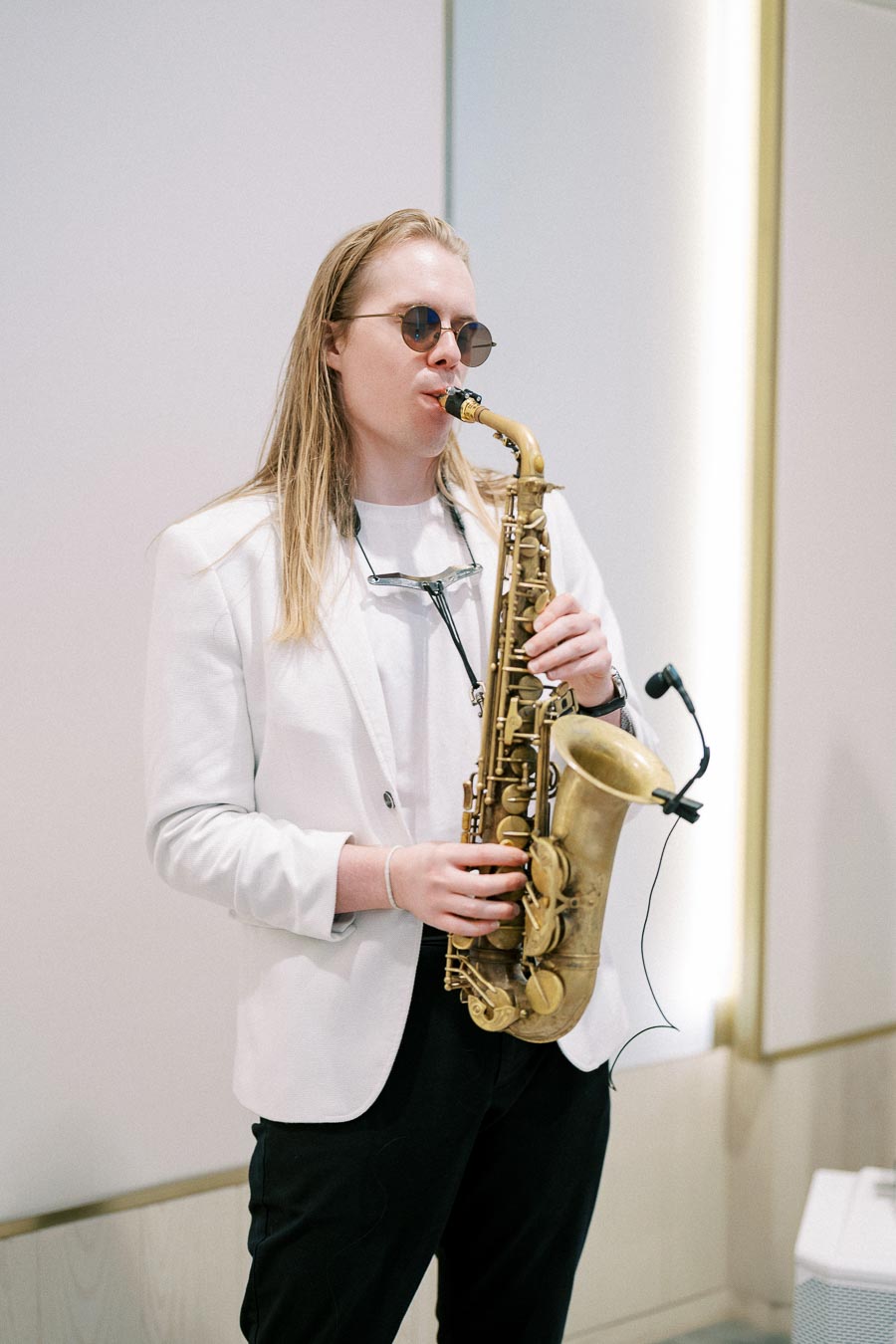 A musician in a white blazer and sunglasses playing a saxophone indoors.