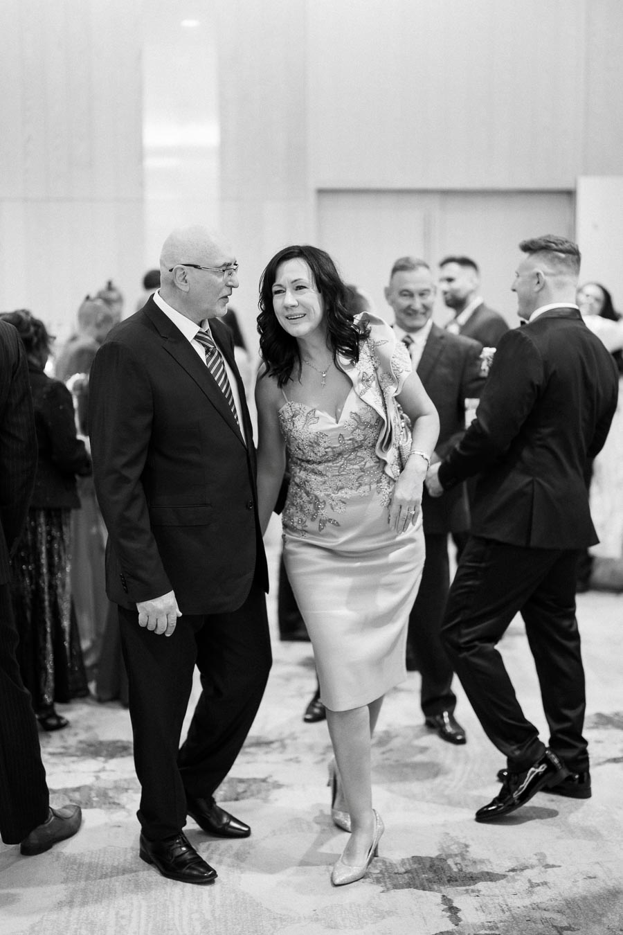 A black and white photo of elegantly dressed people socializing at a formal event, with a woman in a detailed dress and a man in a suit engaged in conversation, surrounded by other guests.