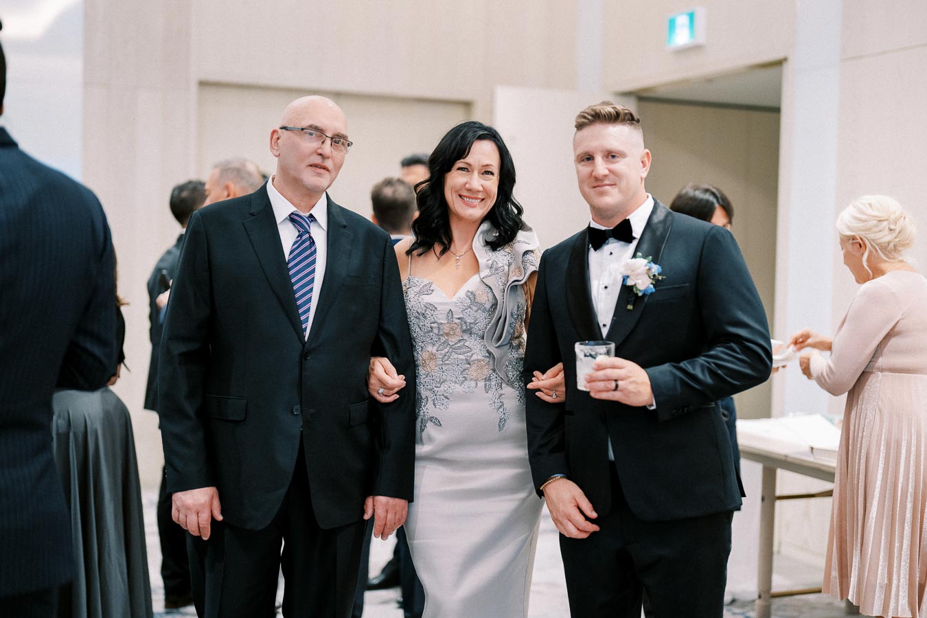 A smiling group of three people at a formal event, with a woman in an elegant gown and two men in suits, one holding a drink.