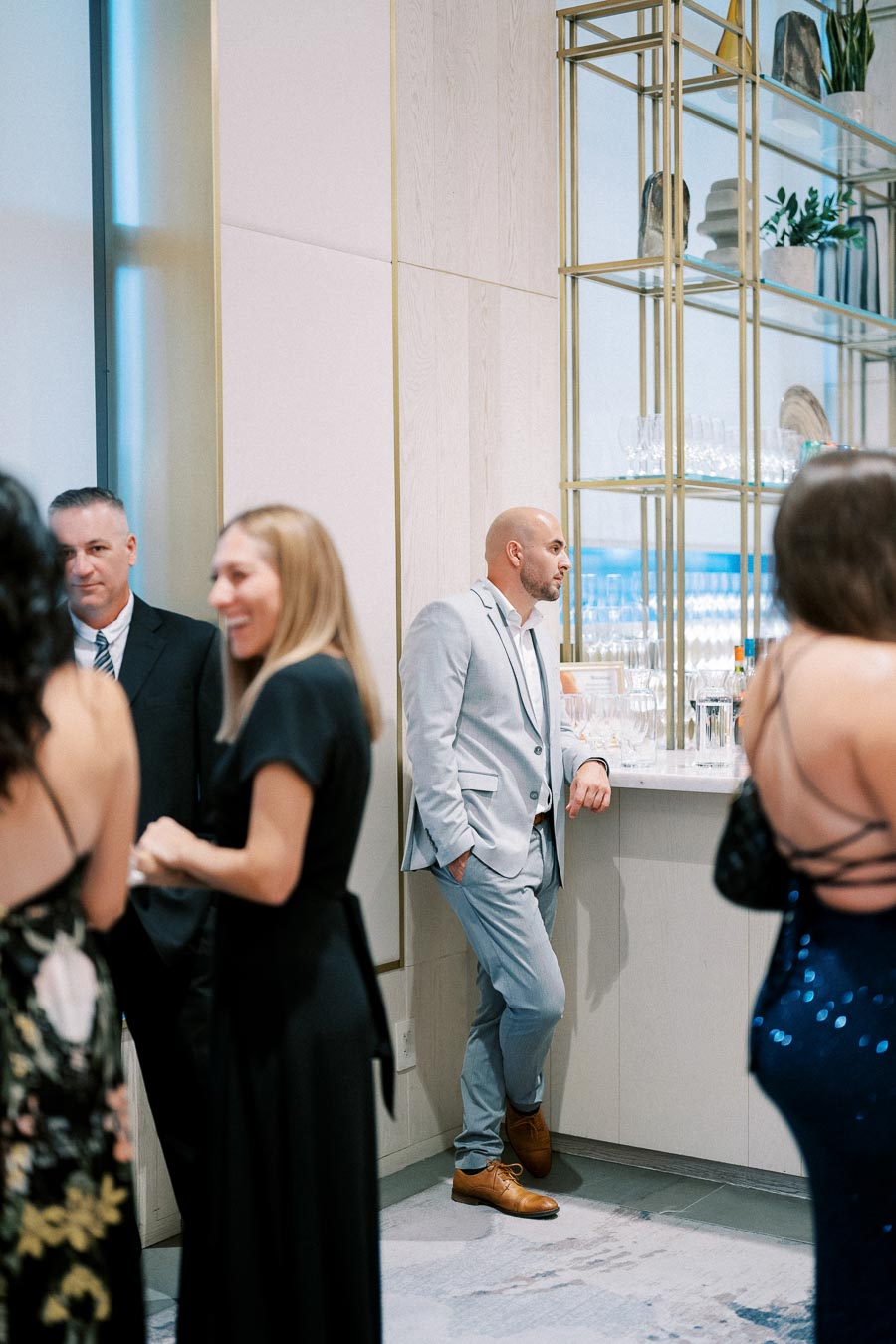 Group of people socializing in an elegant setting with a man in a light suit leaning against a bar.
