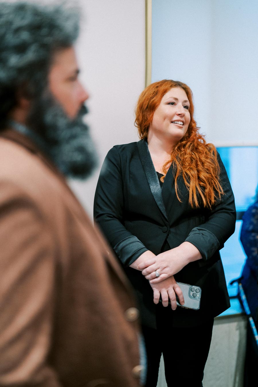 A woman with long red hair in a black blazer smiling during a social gathering. She is holding a smartphone and appears to be engaged in conversation, with a blurred figure in the foreground.