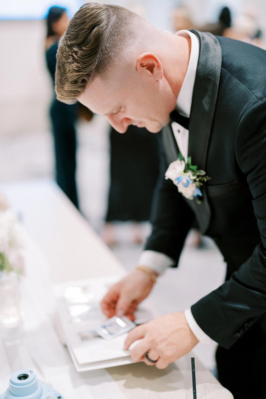 Man in formal suit placing a photo into a guestbook at a wedding reception, with a blue instant camera and drink on the table.