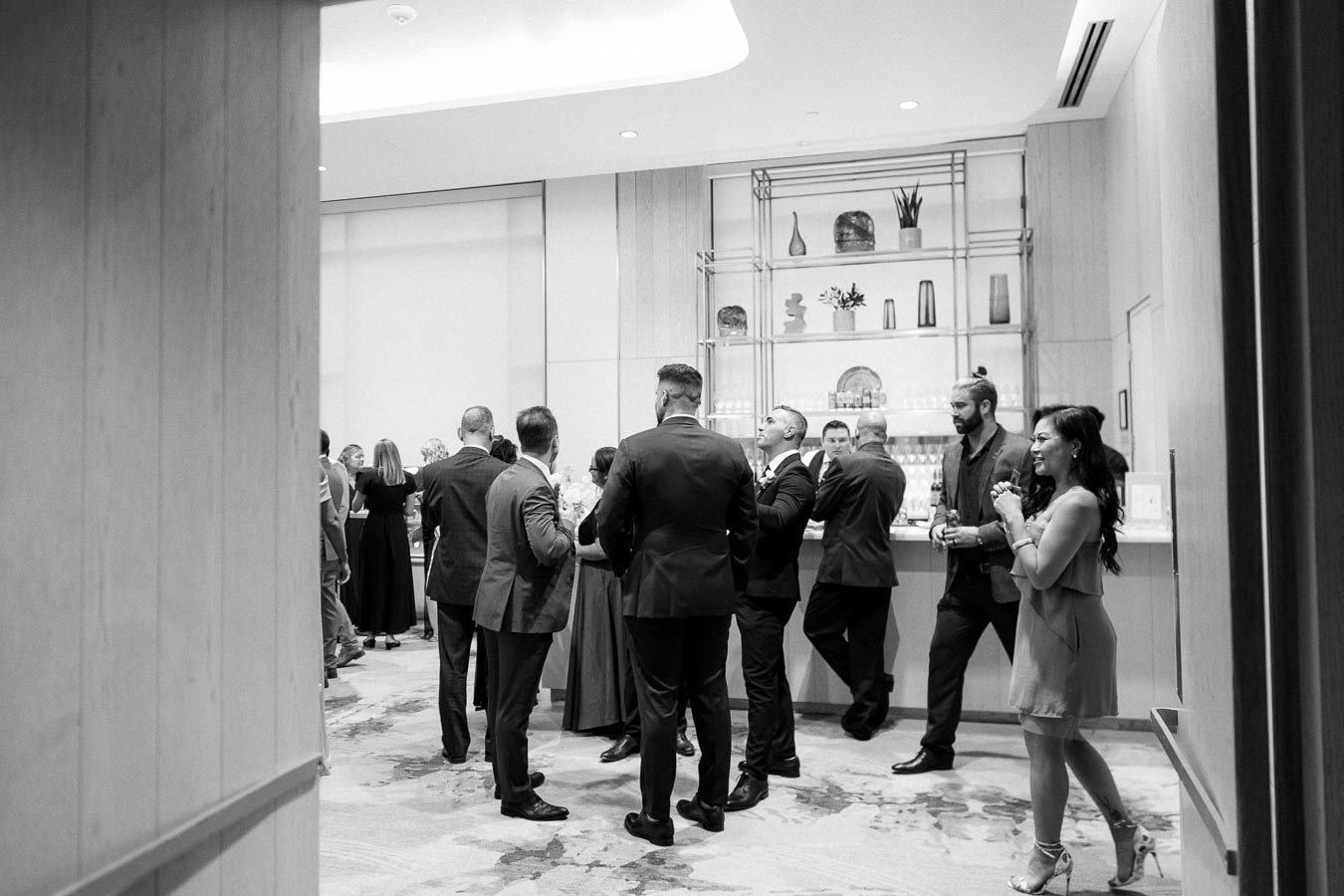 Black and white photo of a group of people dressed formally, engaged in conversation at a stylish indoor event with a decorative shelf and bar in the background.