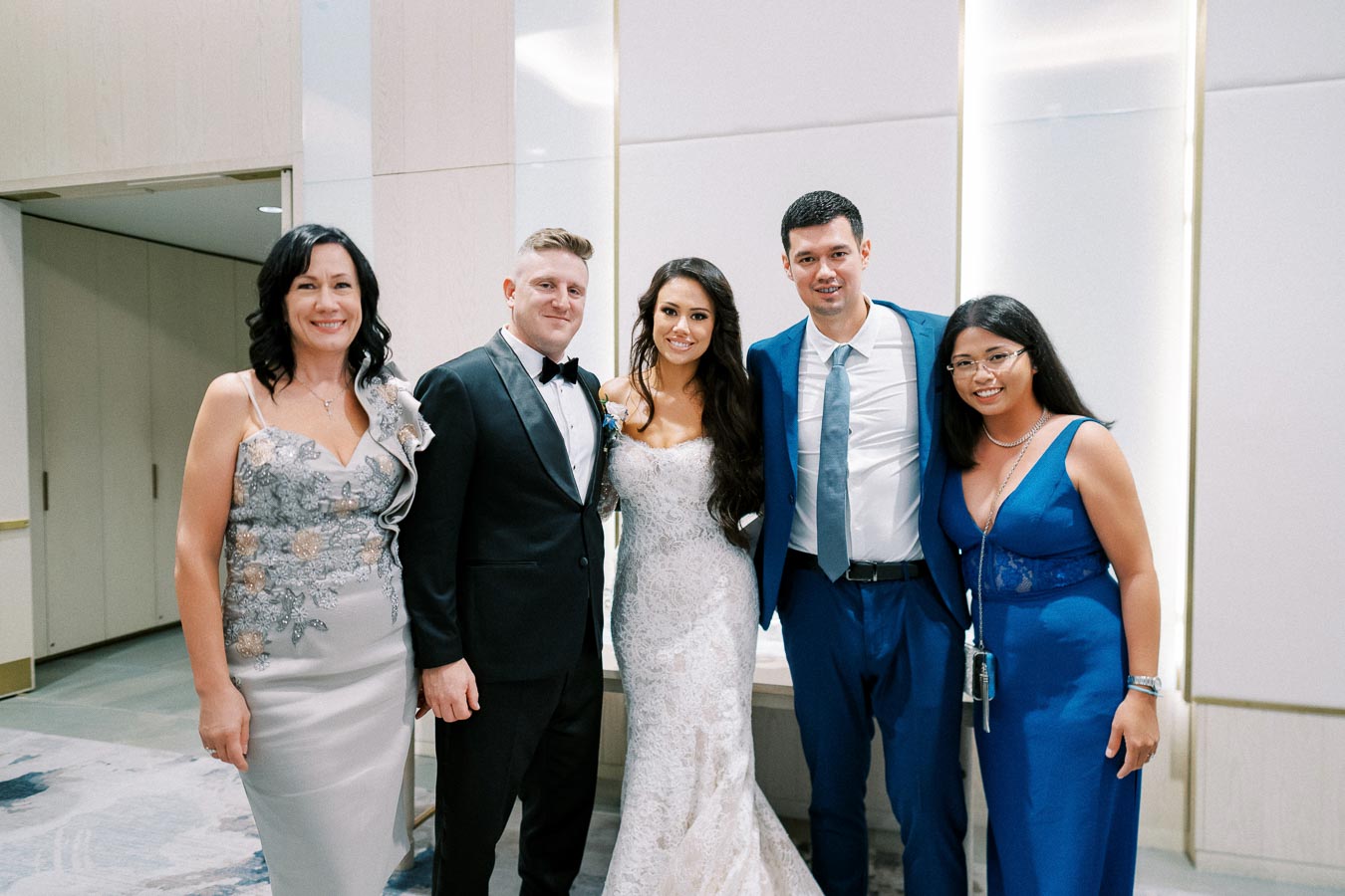 A group of five people dressed in formal attire, including a bride in a white wedding gown, posing together indoors at a wedding reception.