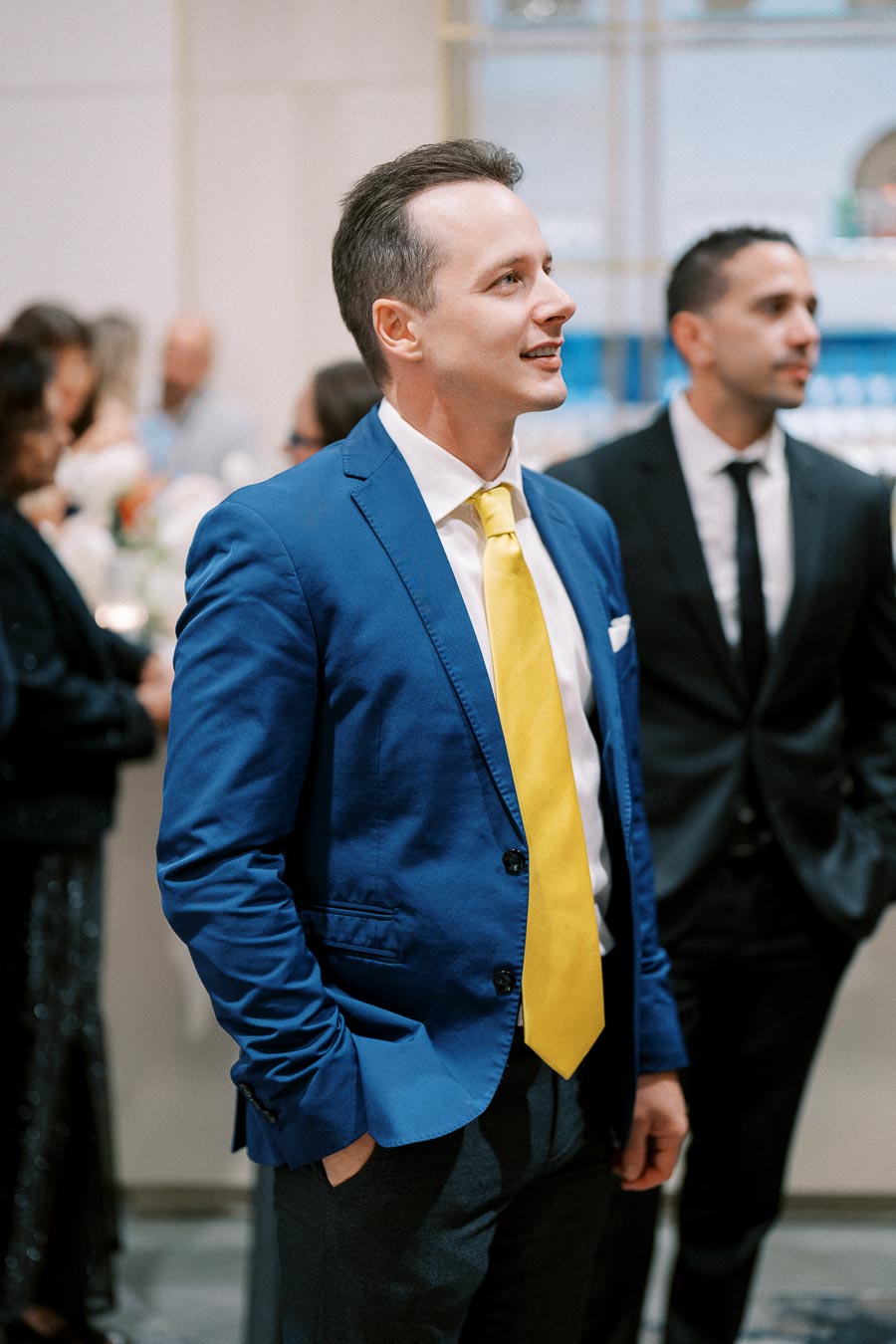 Man in a blue suit and yellow tie at a formal event, surrounded by people in semi-formal attire, engaging in conversation.
