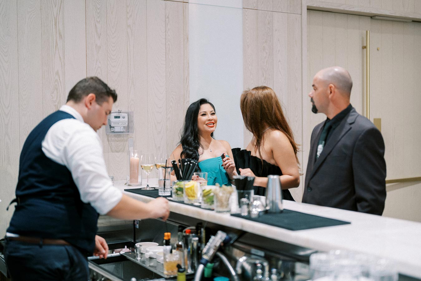 A group of people enjoying drinks and conversation at a stylish bar setting, with a bartender preparing beverages in the foreground.
