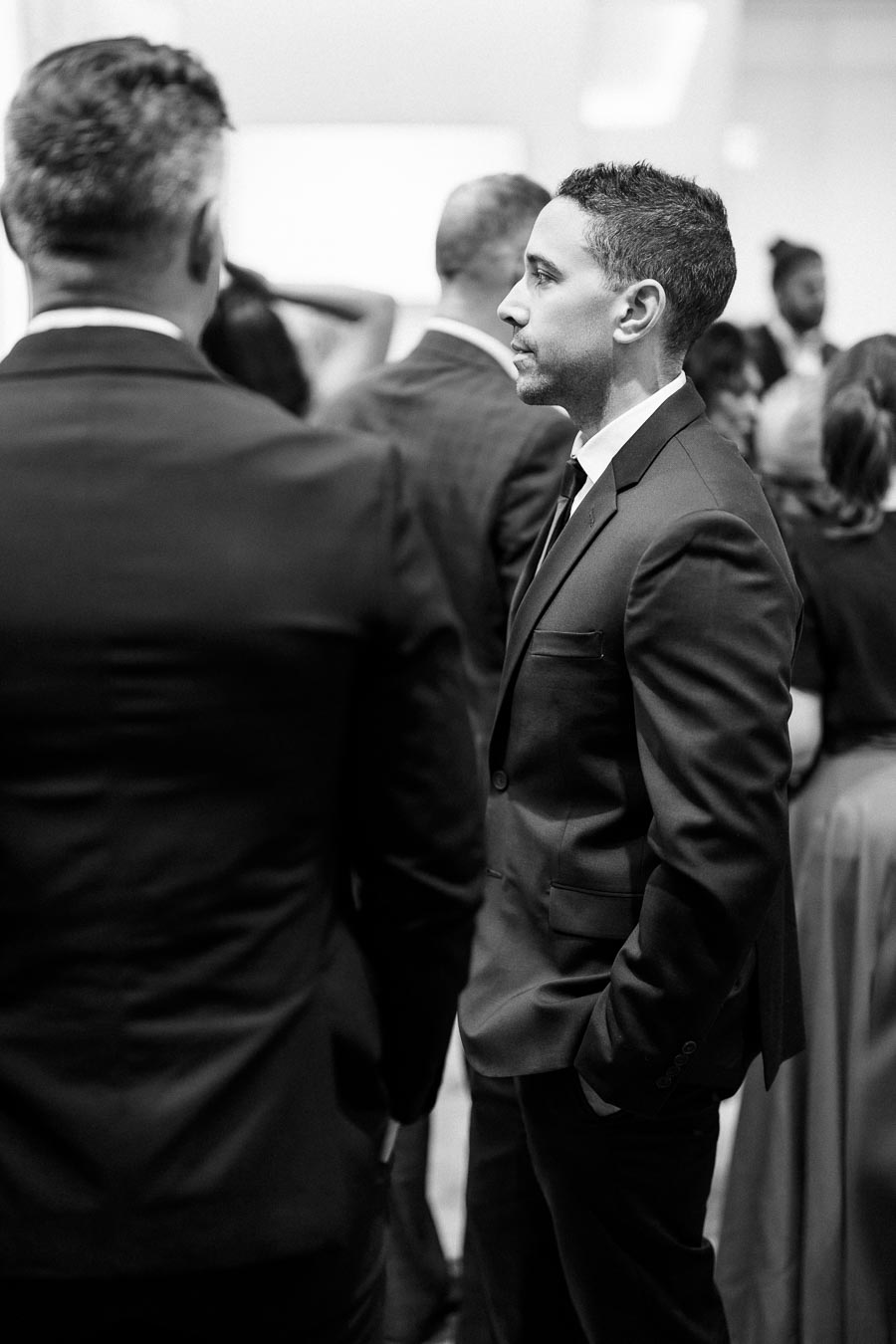 Black-and-white photo of a man in a suit at a formal event, standing amongst a group of people in elegant attire.