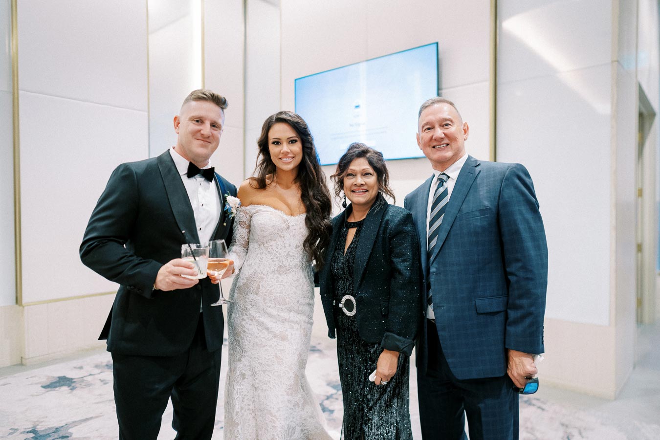 A wedding couple and two guests elegantly dressed in formal attire, smiling at the camera during a celebration in a modern venue. The groom holds a wine glass, and the bride wears a white lace gown.