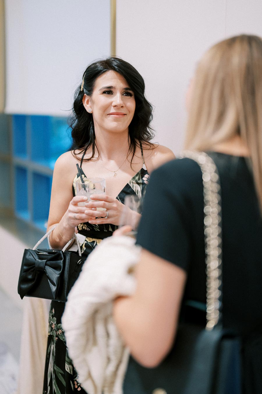 Woman with a cocktail smiling during a social gathering, wearing a floral dress and holding a black handbag.