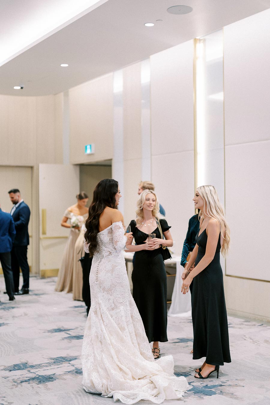 A bride in a lace wedding gown chatting with guests in formal attire during a modern indoor wedding reception.
