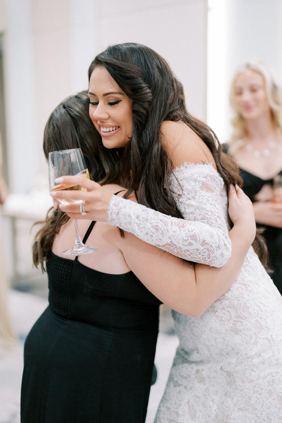 Bride in lace wedding dress hugs guest holding wine glass at wedding reception, showing joy and celebration.