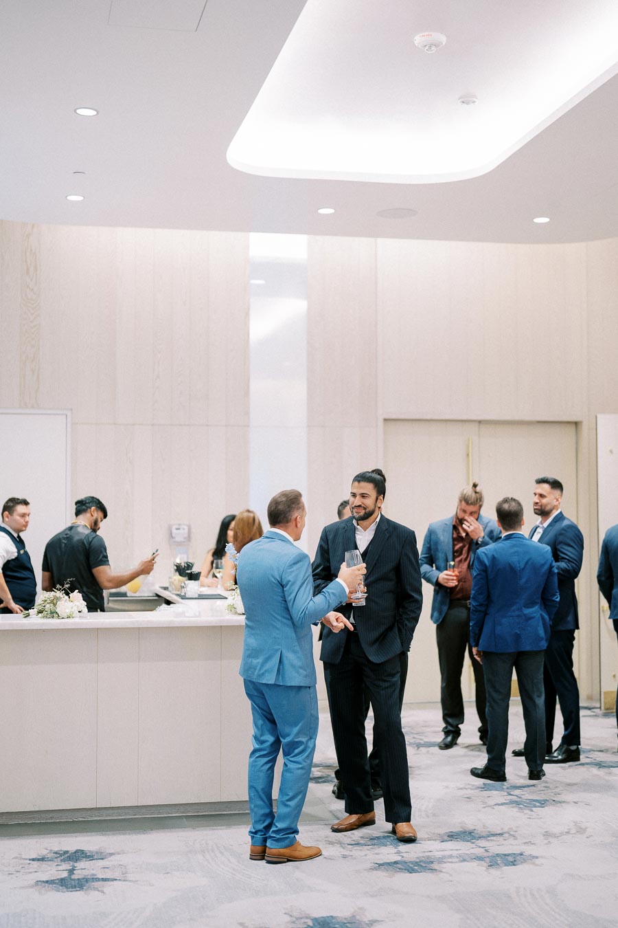A group of business professionals socializing in a modern conference hall, dressed in formal attire, with a bar in the background.