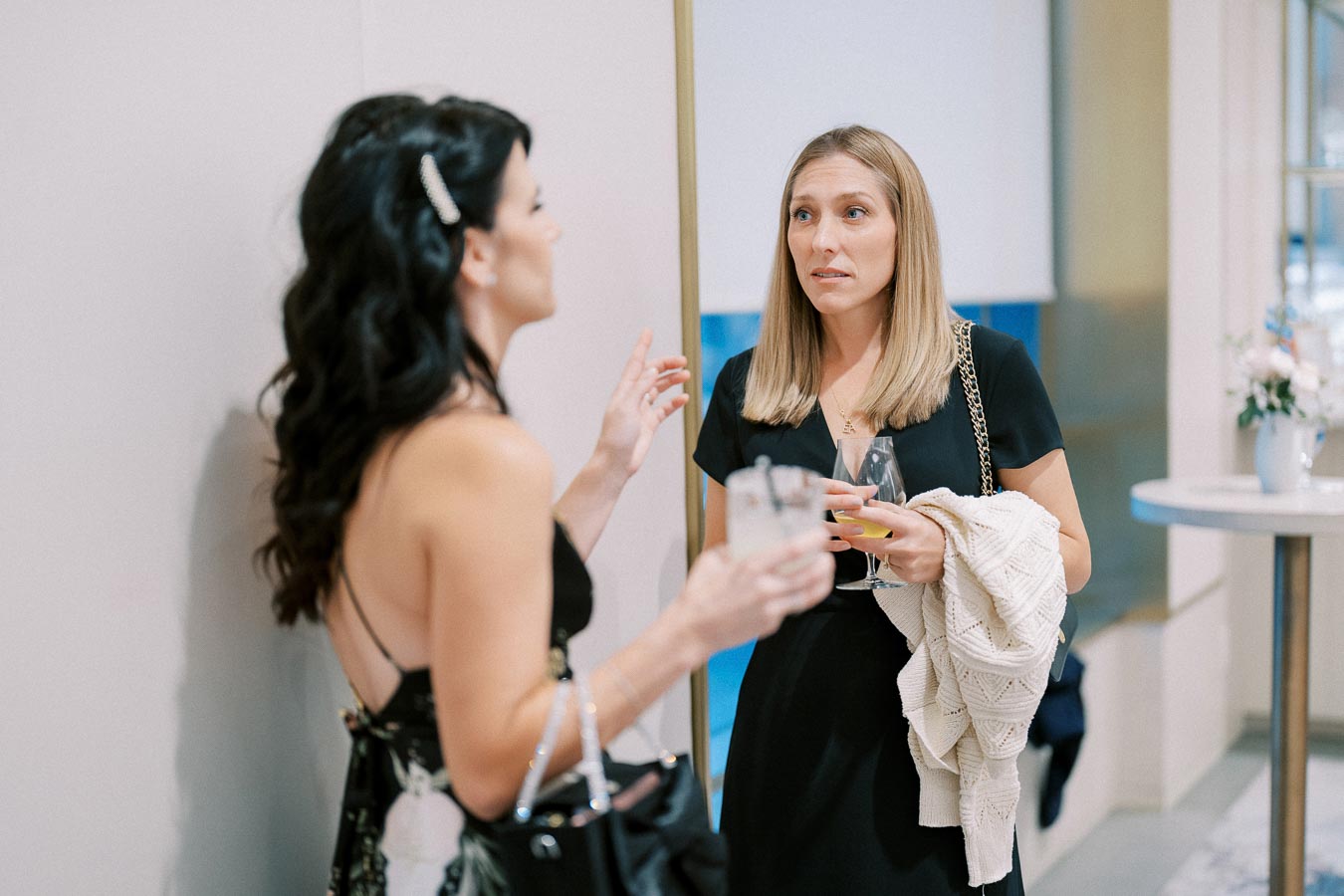 Two women in elegant black dresses engaging in conversation at a social event, each holding a drink in their hands.