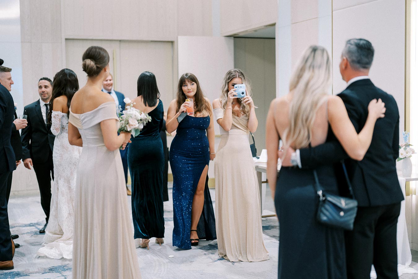 Alt text: Group of elegantly dressed people mingling at a formal event, with women in gowns and men in suits, capturing photos and holding drinks.