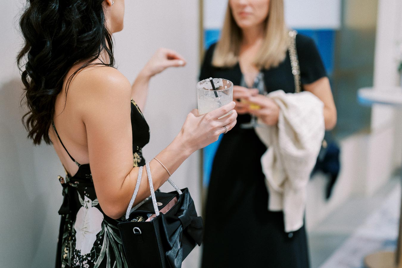 Two women in elegant dresses engage in conversation at a social event, each holding a cocktail in hand.