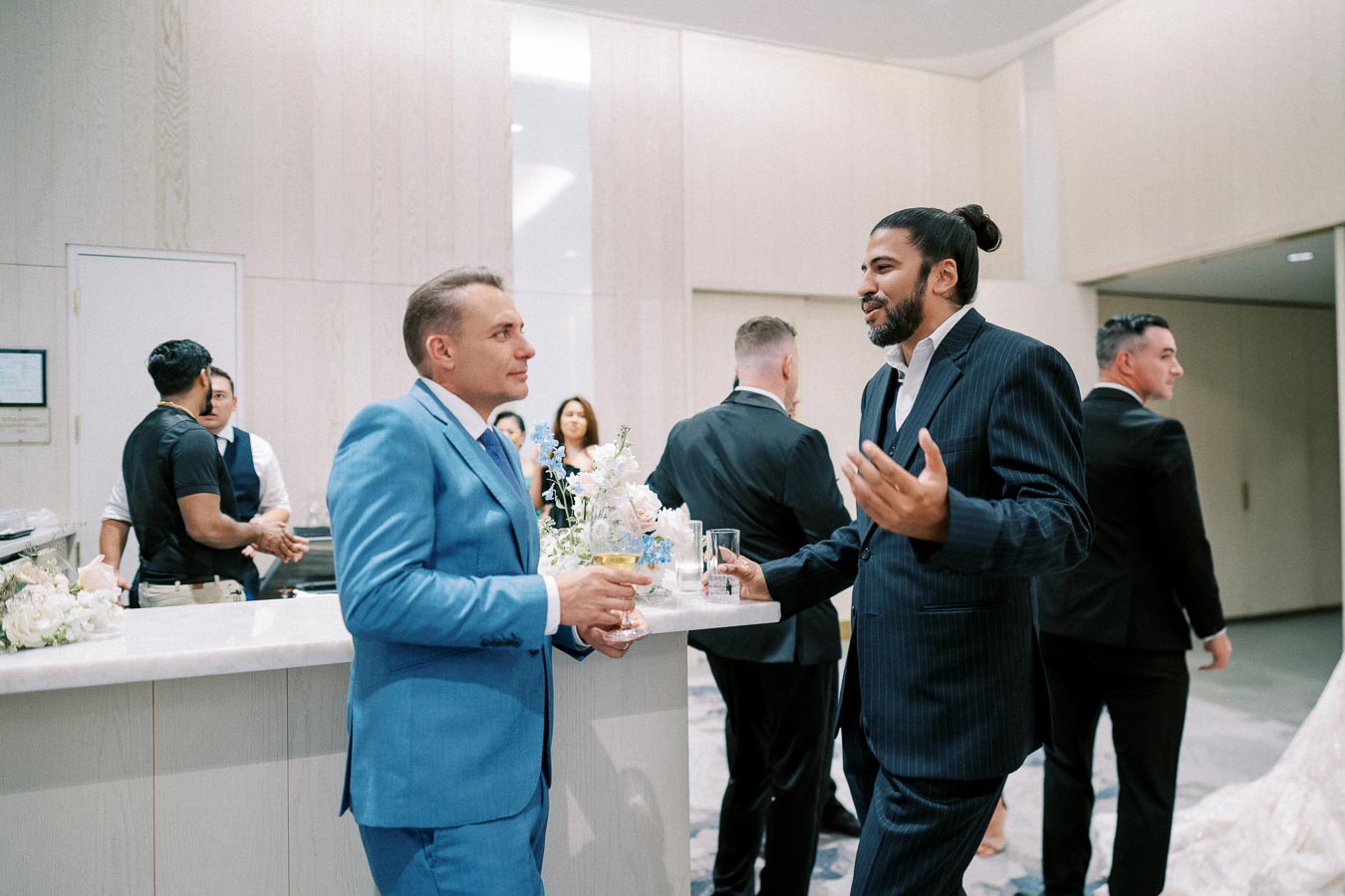Business professionals networking at an event, with a man in a blue suit conversing with a man in a dark suit near a countertop adorned with floral arrangements.