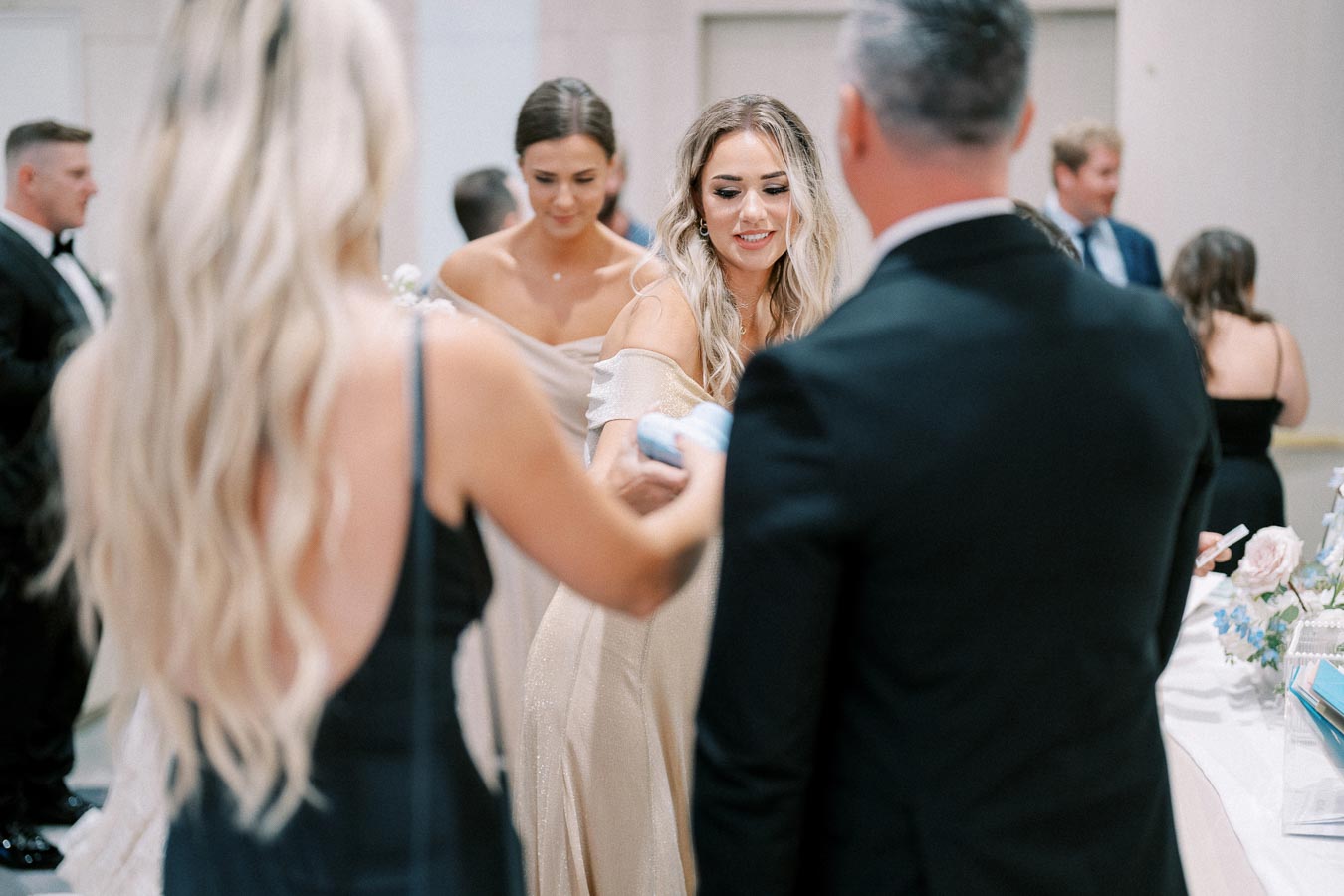 Elegant wedding reception scene with guests in formal attire, including a woman in a shimmering gold dress, socializing around a decorated table.