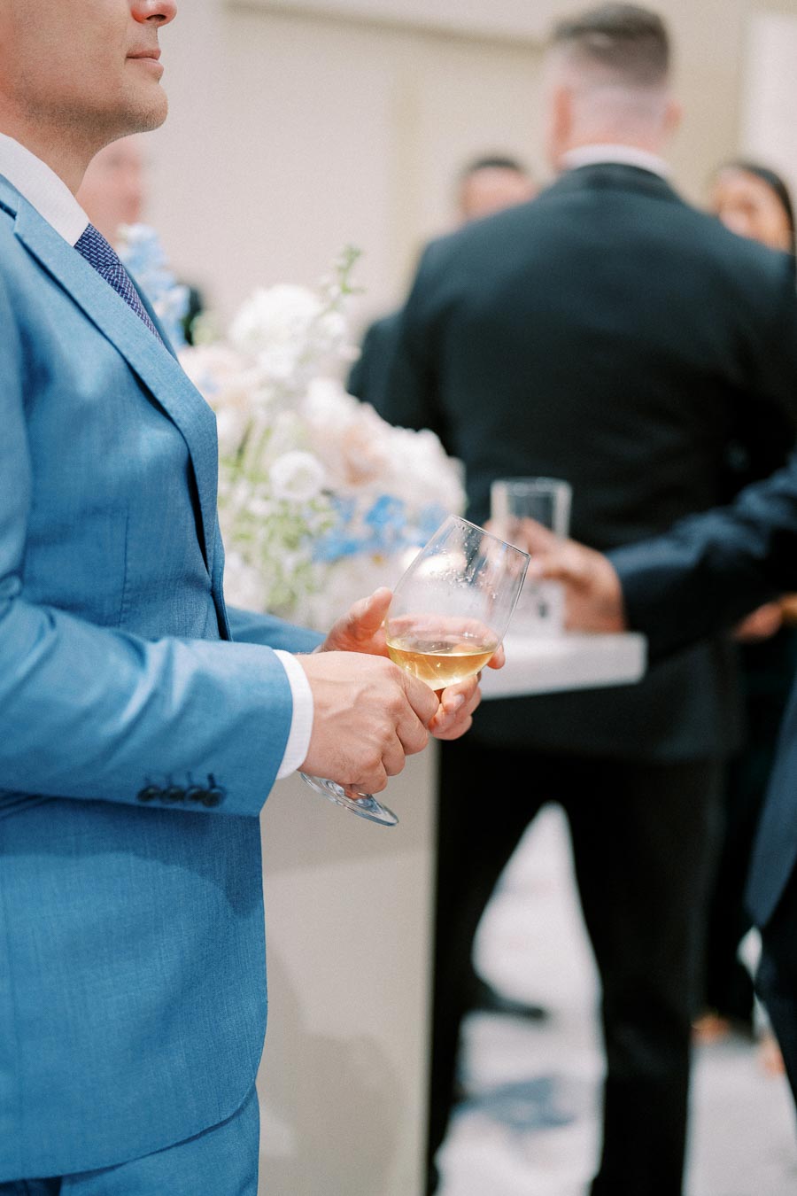 Man in a blue suit holding a glass of white wine at a formal event gathering with blurred guests in the background.