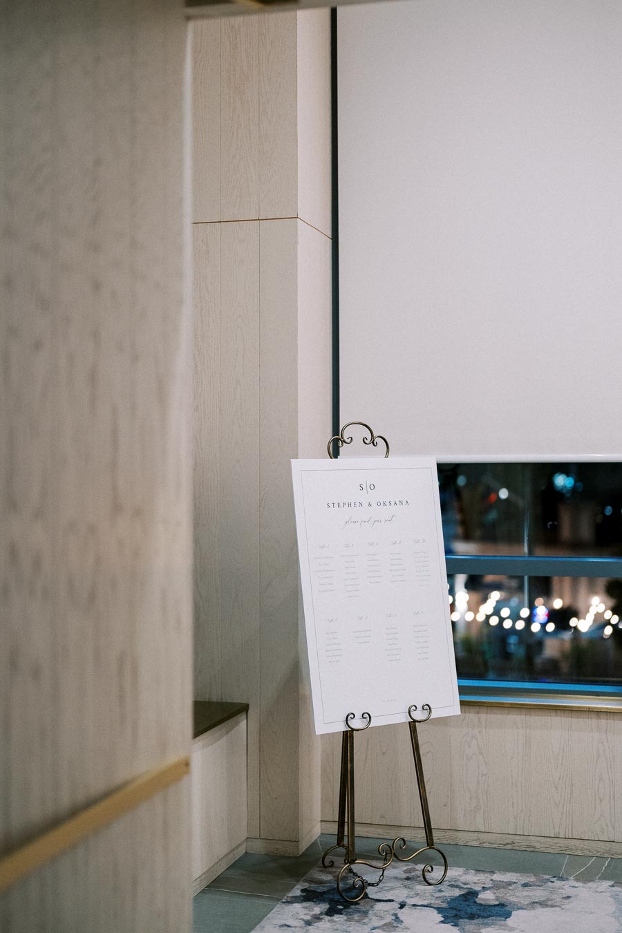Walkway with decorative board on an easel displaying wedding seating chart, set against a modern interior with wooden paneling and a large window.