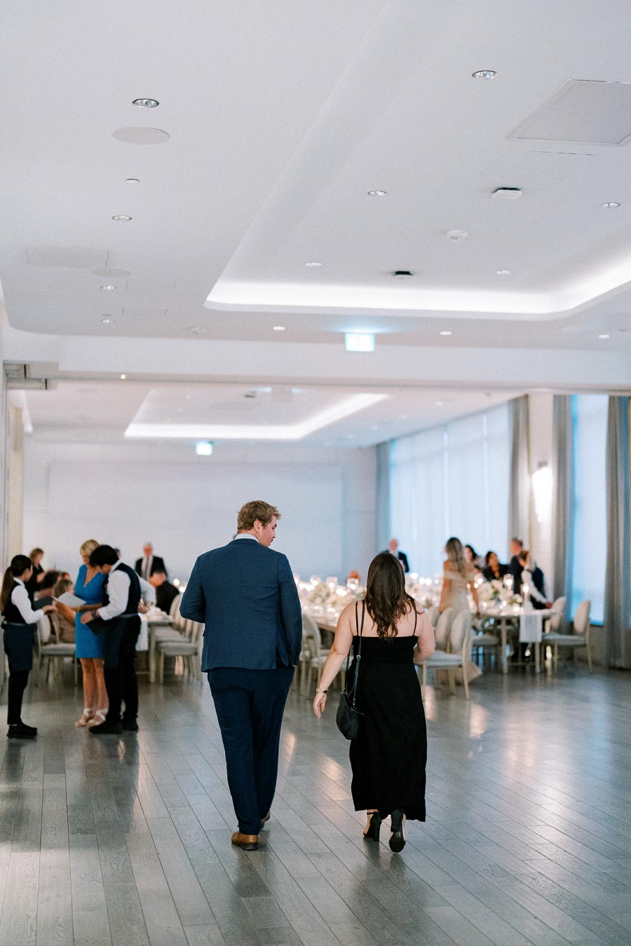 A couple walks towards a dining area in an elegantly decorated venue, filled with guests seated at tables adorned with candles and flowers, during a formal event or wedding reception.